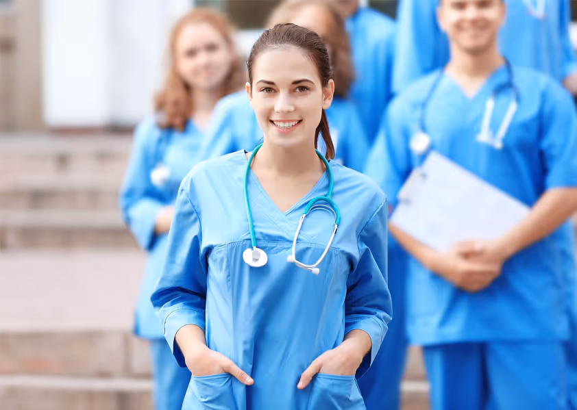 Smiling healthcare professional in blue scrubs with stethoscope standing in front of colleagues outdoors.