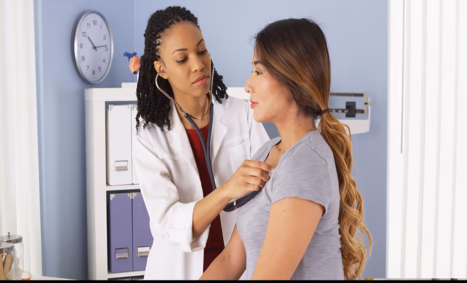 Female doctor using a stethoscope to listen to a female patient’s chest in a medical office.