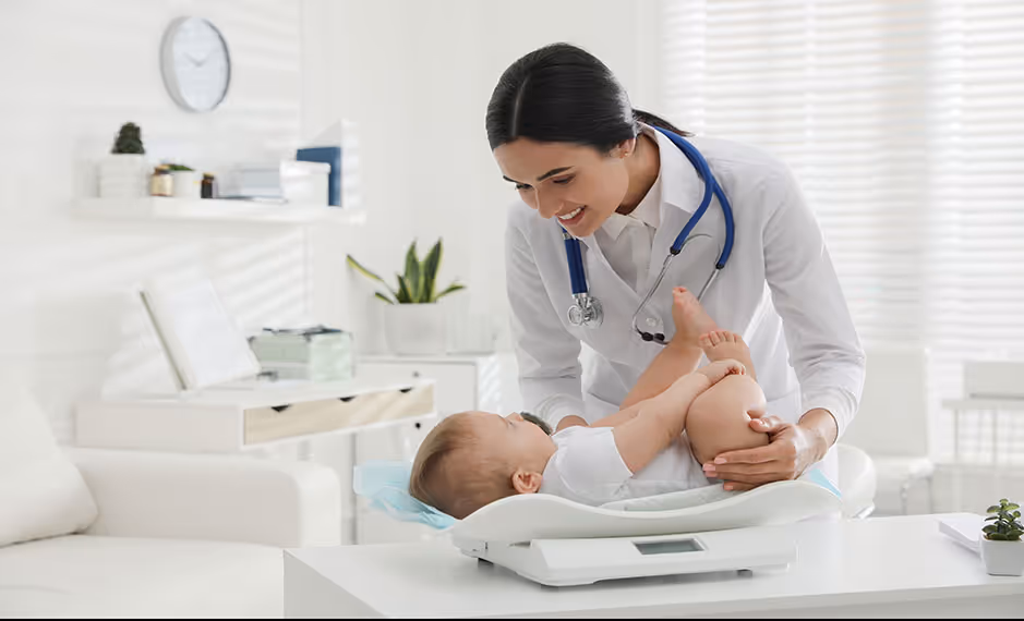 Female doctor with stethoscope smiling at a baby lying on a scale in a bright medical office.