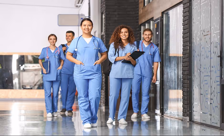 Group of smiling healthcare professionals walking down a hospital corridor in blue scrubs.