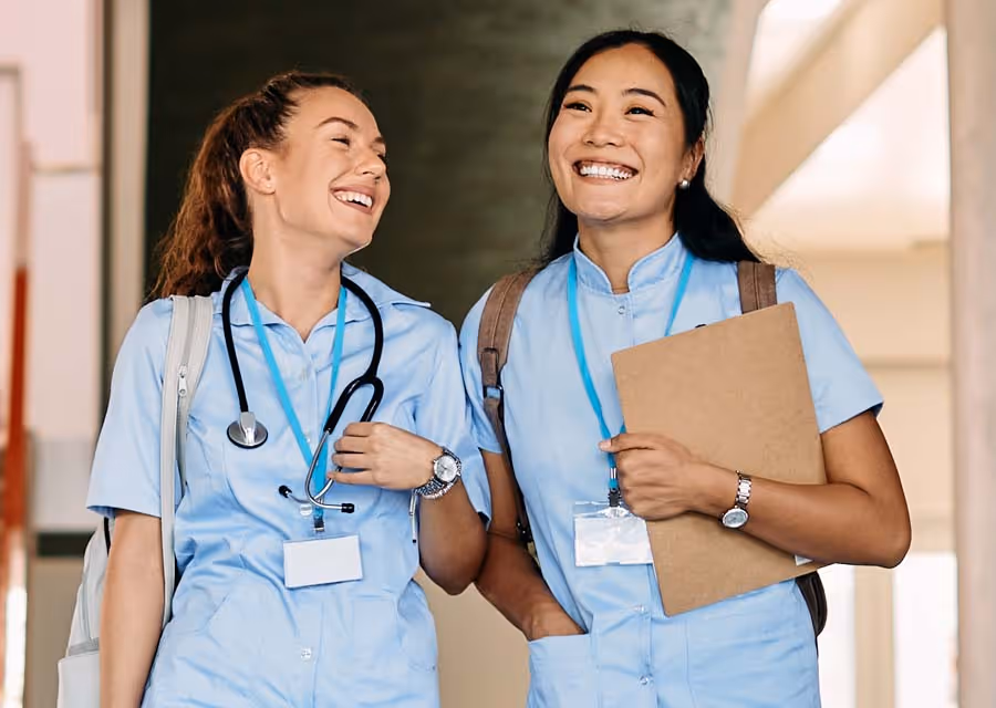 Two smiling female healthcare workers walking indoors, one with stethoscope, one holding clipboard.