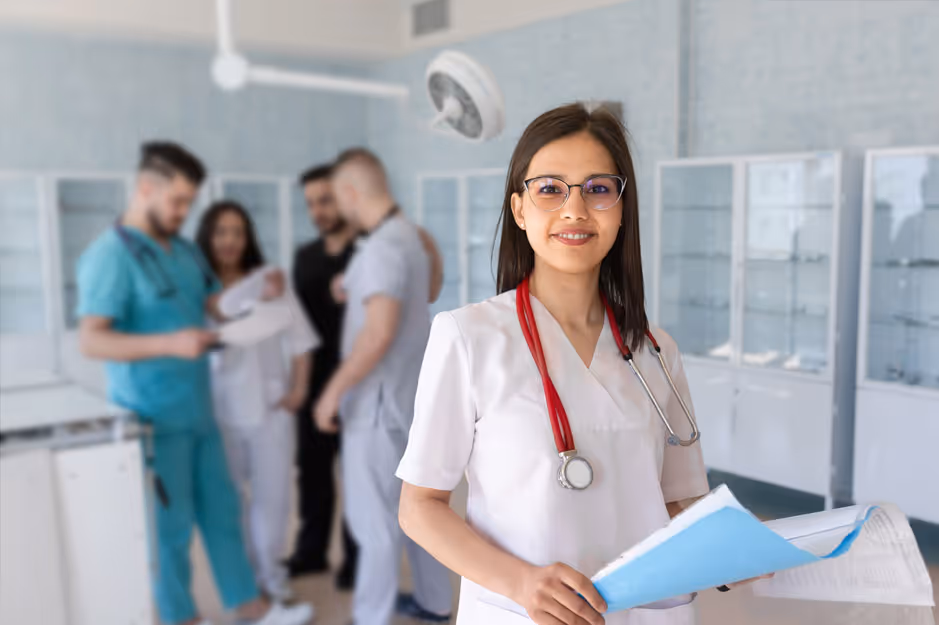 Smiling female medical professional in white scrubs holding files with colleagues in the background.