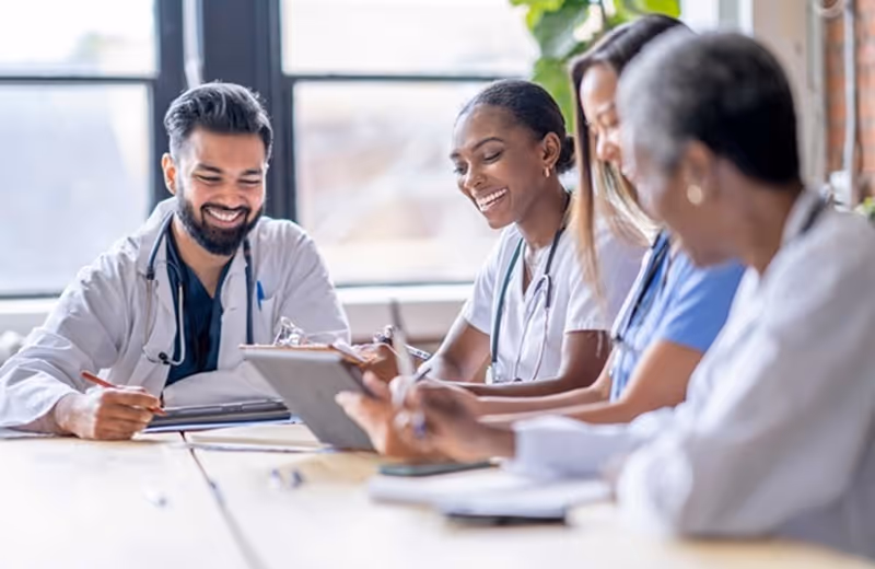 Four diverse healthcare professionals smiling and discussing together around a table in a bright room.