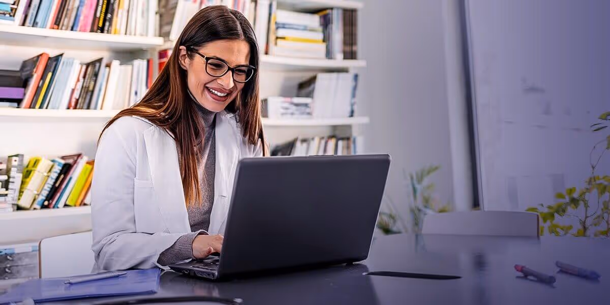 Smiling woman in glasses and white coat working on laptop at desk with bookshelves in background.