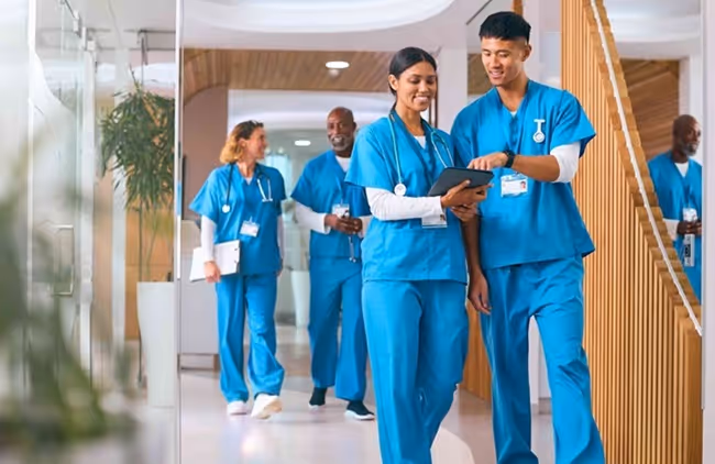 Four healthcare workers in blue scrubs walking and discussing in a modern hospital hallway.