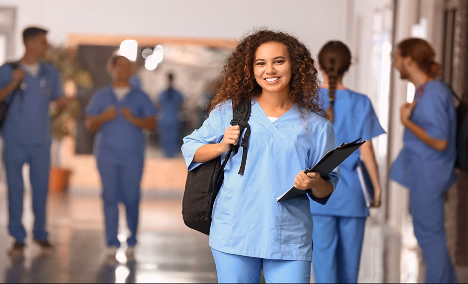 Smiling nurse in blue scrubs carrying backpack and clipboard walking in hospital hallway with colleagues.