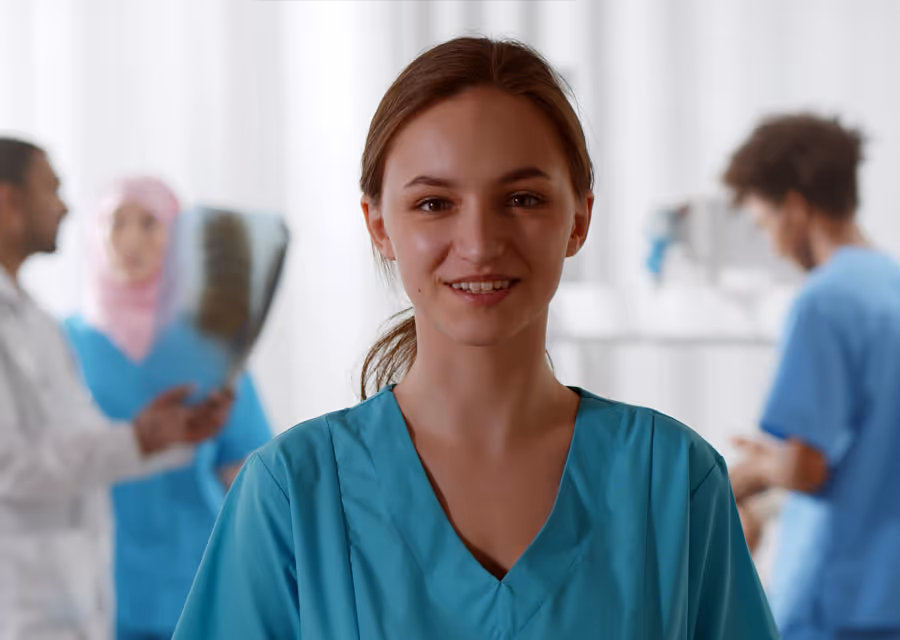 Smiling female nurse in blue scrubs with medical staff talking and examining x-ray behind her.