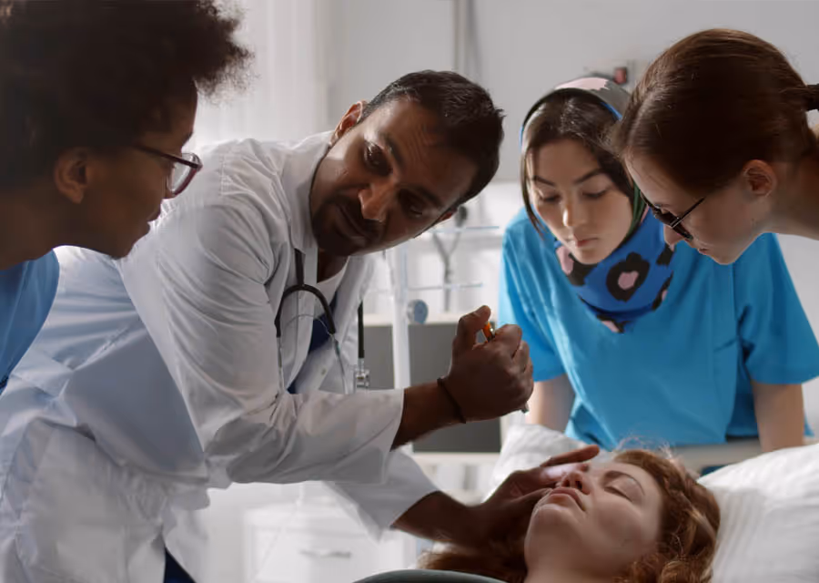 Doctor demonstrates injection technique to three medical students around a patient in bed.