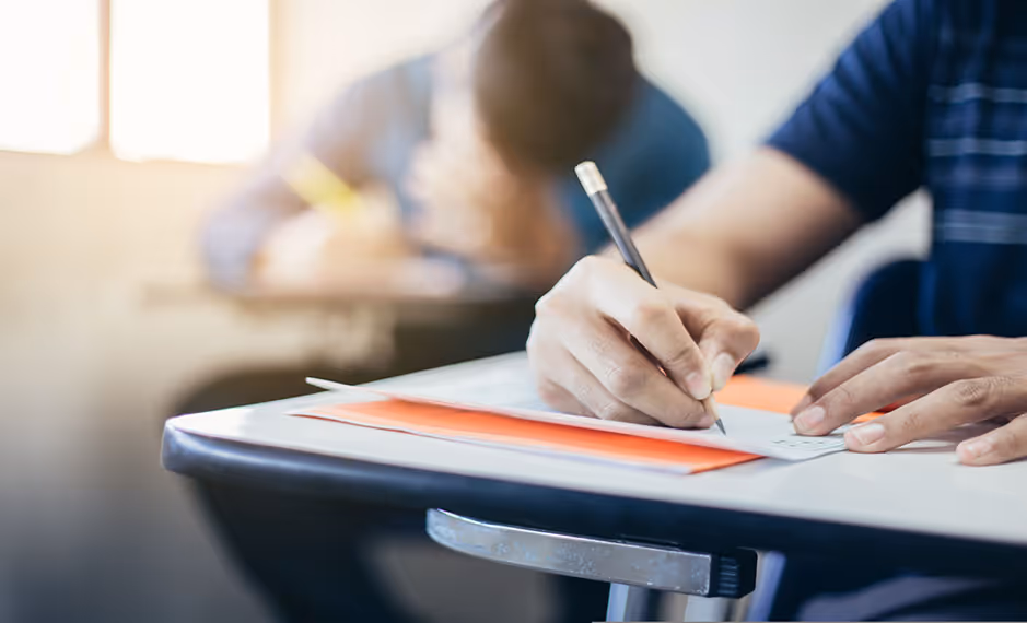 Student holding pencil and completing a test paper at a classroom desk during an exam.