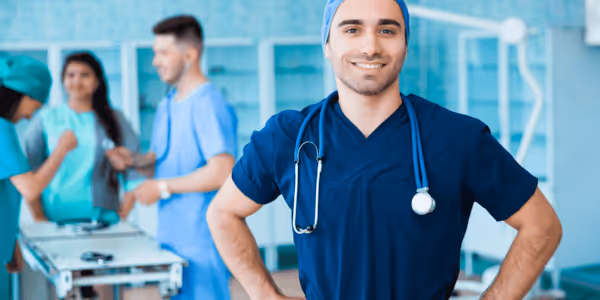 Smiling male nurse in blue scrubs with stethoscope stands in hospital room with colleagues in background.