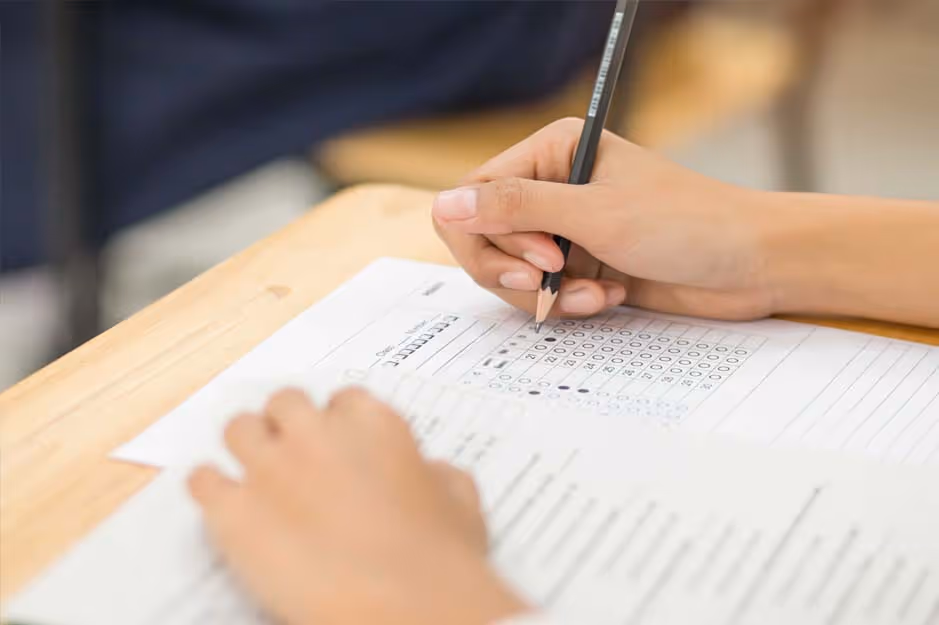 Student filling in multiple-choice answer sheet with pencil on wooden desk during exam.