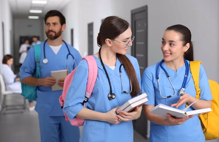 Two female medical students in blue scrubs chatting with books, male student behind in a hallway.