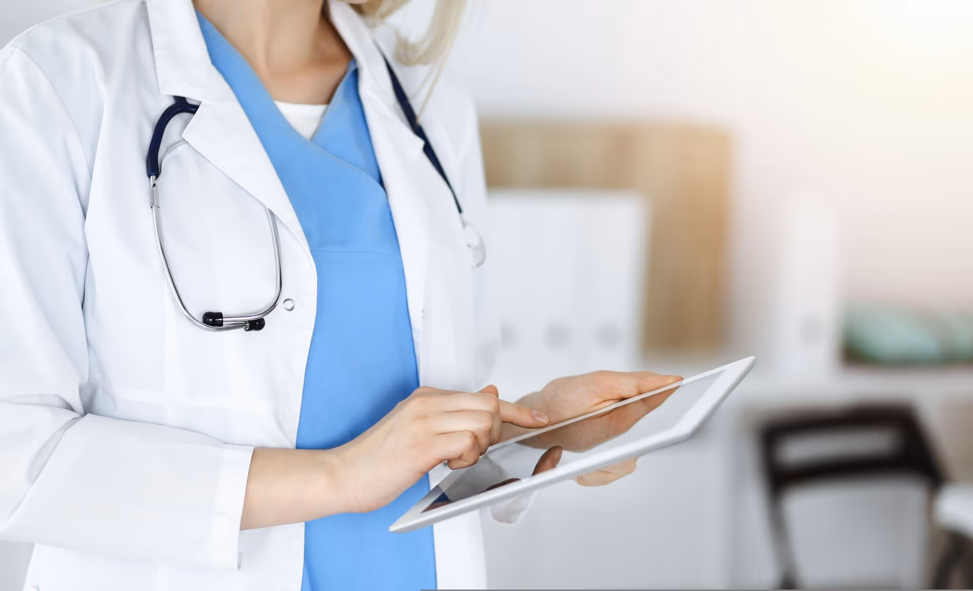 Medical professional in blue scrubs and white coat using a tablet with a stethoscope around the neck.