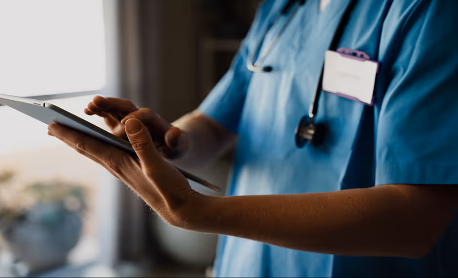 Healthcare worker in blue scrubs using a tablet, with a stethoscope around their neck.