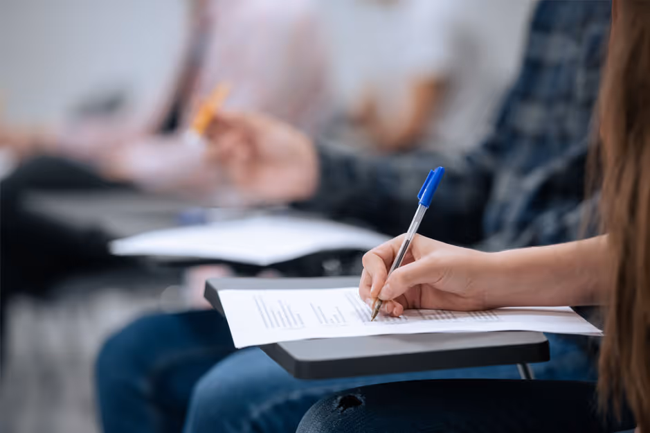 Student writing with a blue pen on paper resting on a desk in a classroom setting.