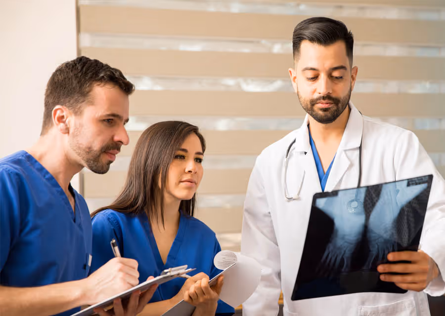 Three medical professionals reviewing a hand x-ray, two in blue scrubs taking notes.