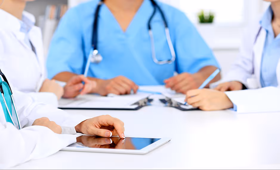 Medical professionals sitting at a table with a tablet, papers, and clipboards during a discussion.