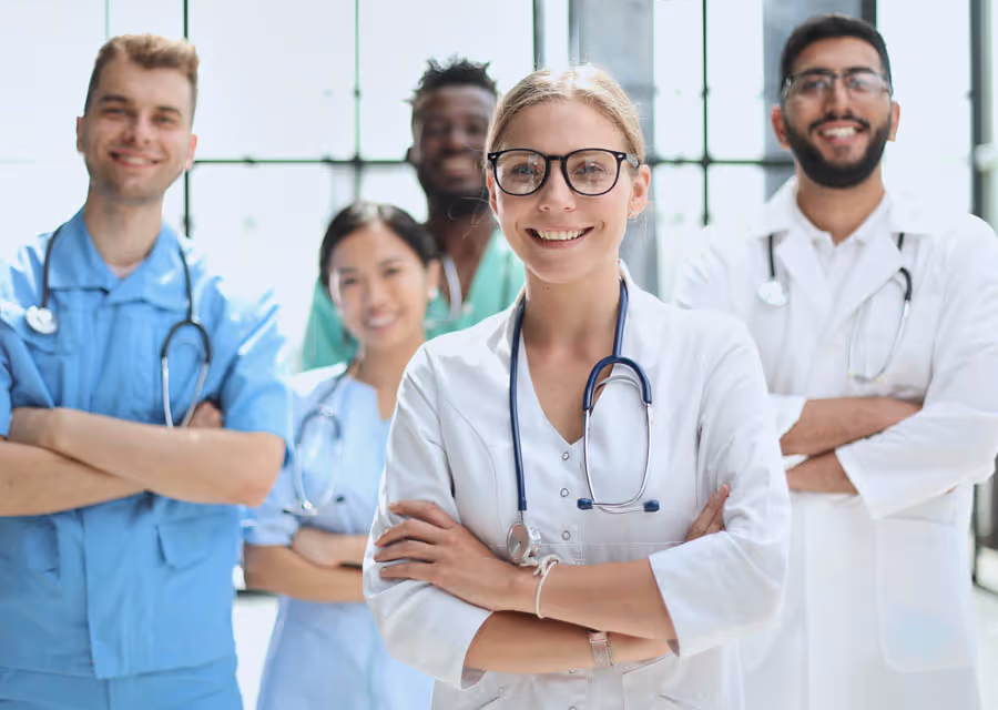 Five diverse medical professionals smiling with arms crossed, wearing scrubs and lab coats with stethoscopes.