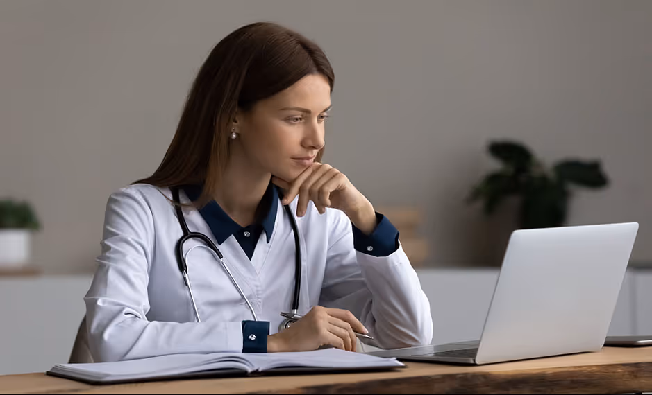 Female doctor in white coat with stethoscope looking at a laptop while sitting at a desk with a notebook.