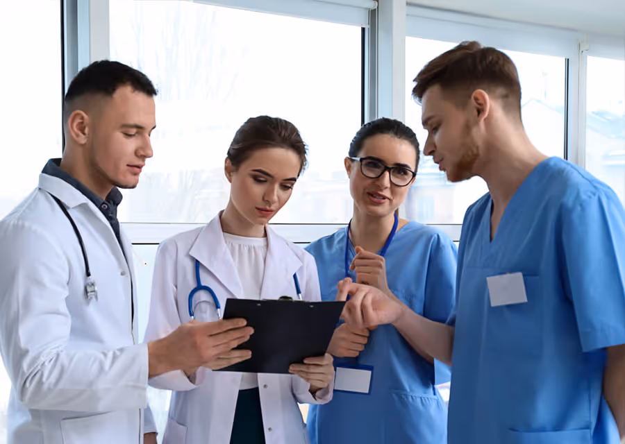 Group of four doctors and nurses reviewing patient info on clipboard in hospital room.