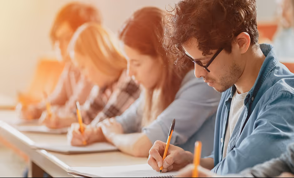 Students sitting in a row, focused and writing in notebooks during a class or exam.