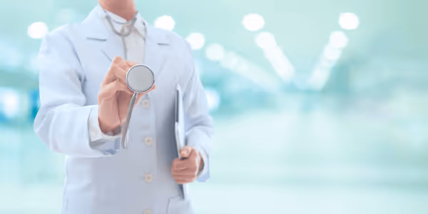Doctor in white coat holding a stethoscope towards the camera in a bright hospital corridor.