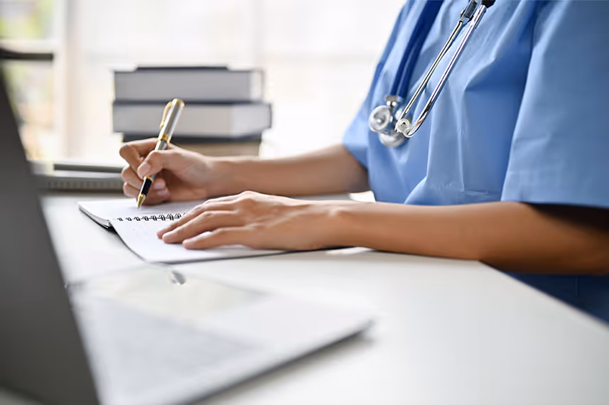 Healthcare professional in blue scrubs writing notes with a stethoscope around their neck.