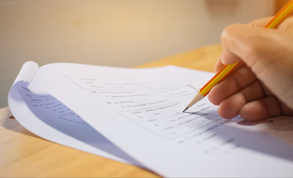 Hand holding a pencil marking answers on a multiple-choice test on a wooden desk.