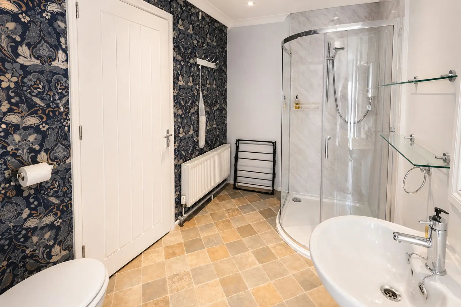 A bright bathroom featuring a shower, basin, and toilet and a decorative blue and silver floral wallpaper accent