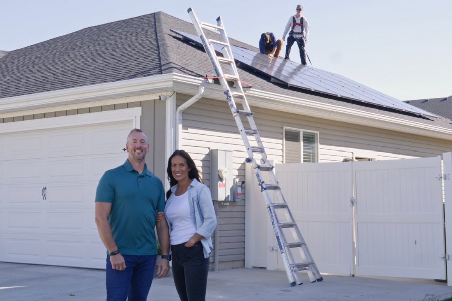 A smiling couple stands outside their garage while Big Dog Solar technicians install solar panels on the roof using a secured ladder and safety gear.