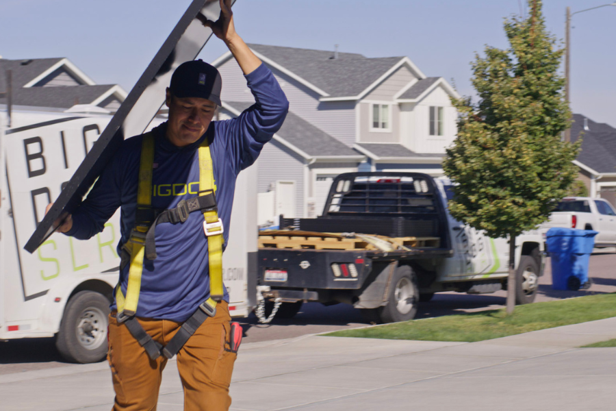 A Big Dog Solar technician carries a solar panel across a residential street, with branded work trucks and equipment visible in the background.