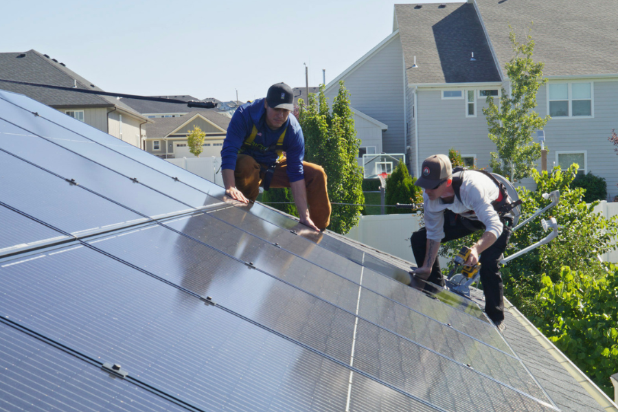 Two solar panel installers work together on a rooftop in a residential neighborhood, carefully aligning and securing panels in bright daylight.
