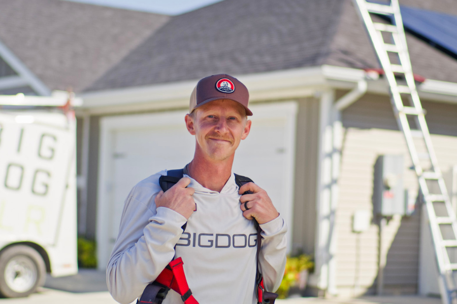 Smiling Big Dog Solar technician in branded gear stands confidently in front of a home with solar panels and ladders visible behind him.