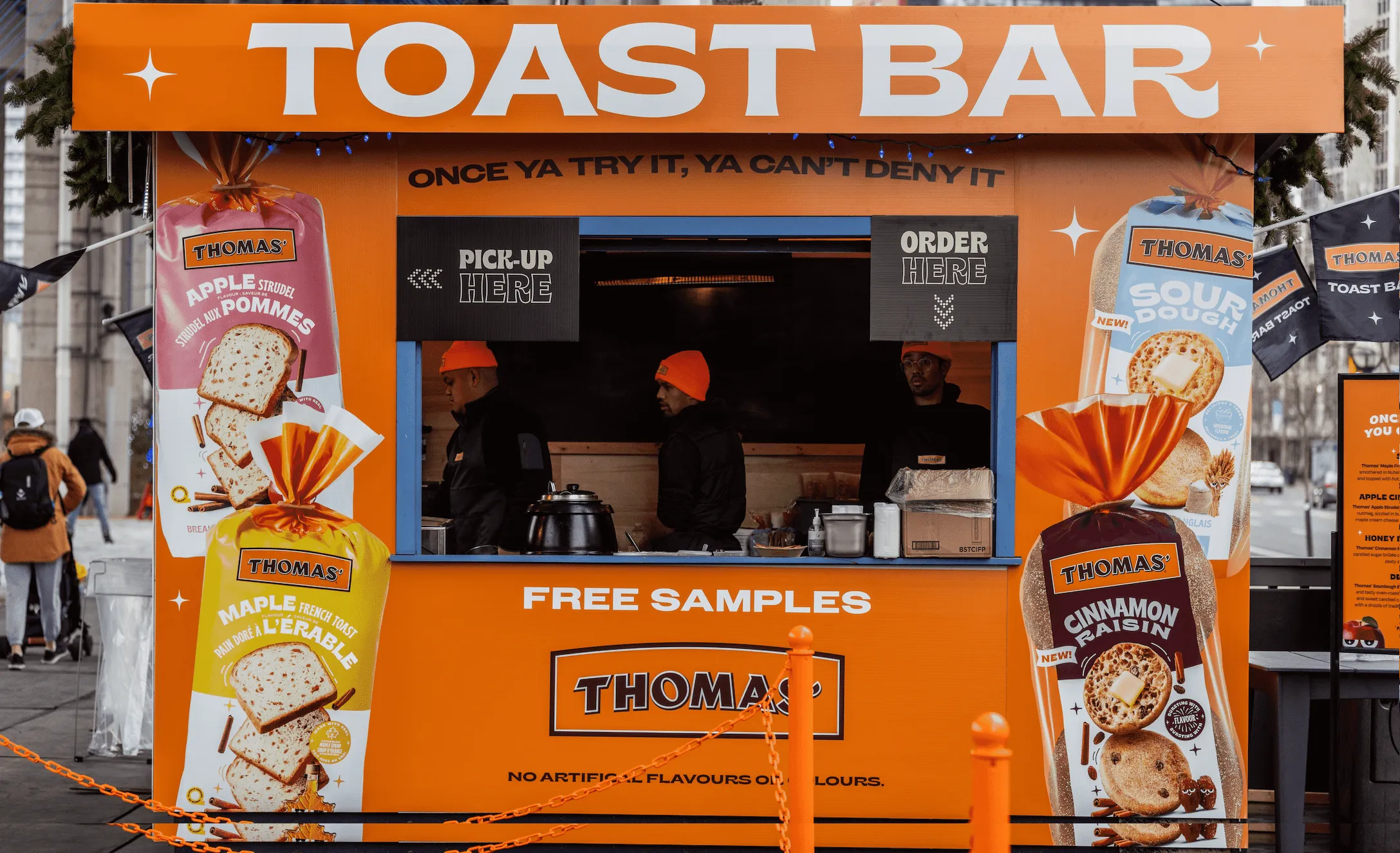 Orange Thomas Toast Bar stand with employees in orange hats serving free samples of various Thomas bread flavors including apple strudel, maple French toast, sourdough, and cinnamon raisin.