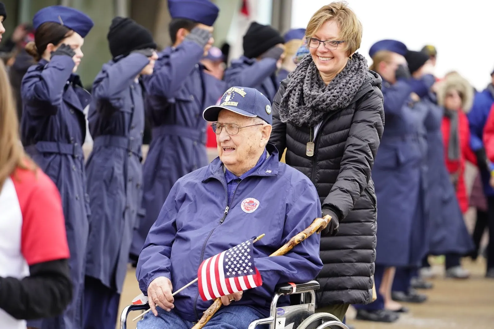 Photograph of a woman pushing a wheelchair where an older veteran sits and holds a U.S. flag and a cane as people salute.