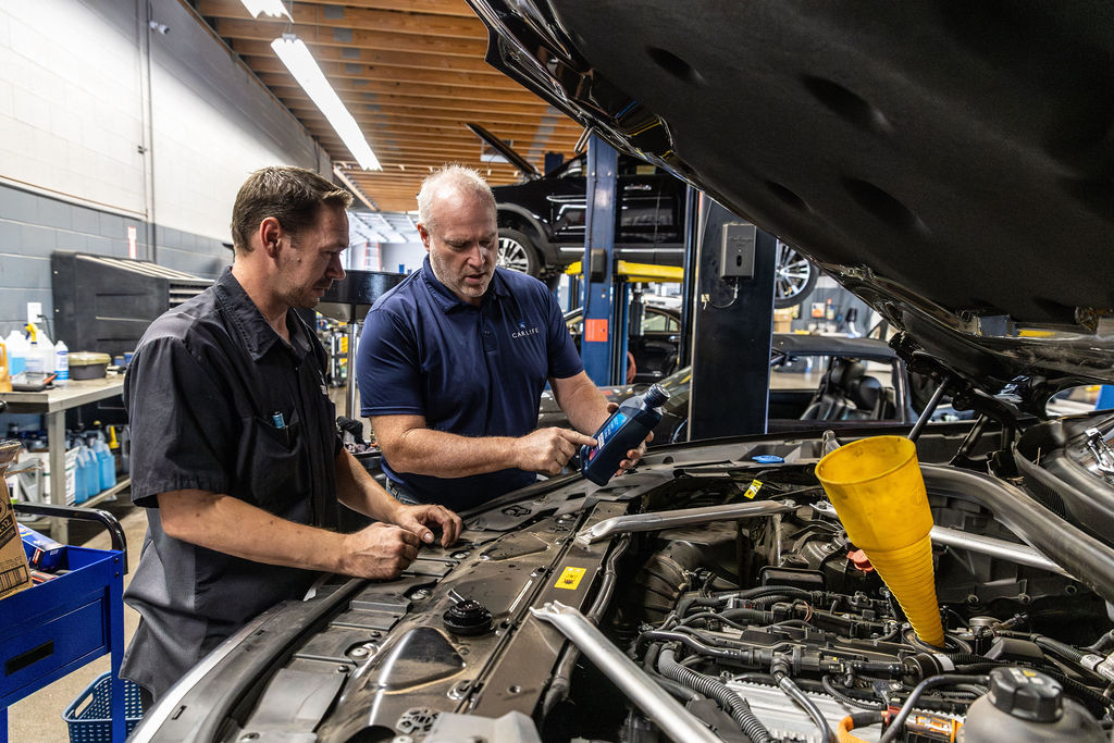 Two mechanics inspecting an engine with one pointing to a bottle of motor oil and a yellow funnel placed in the engine bay.