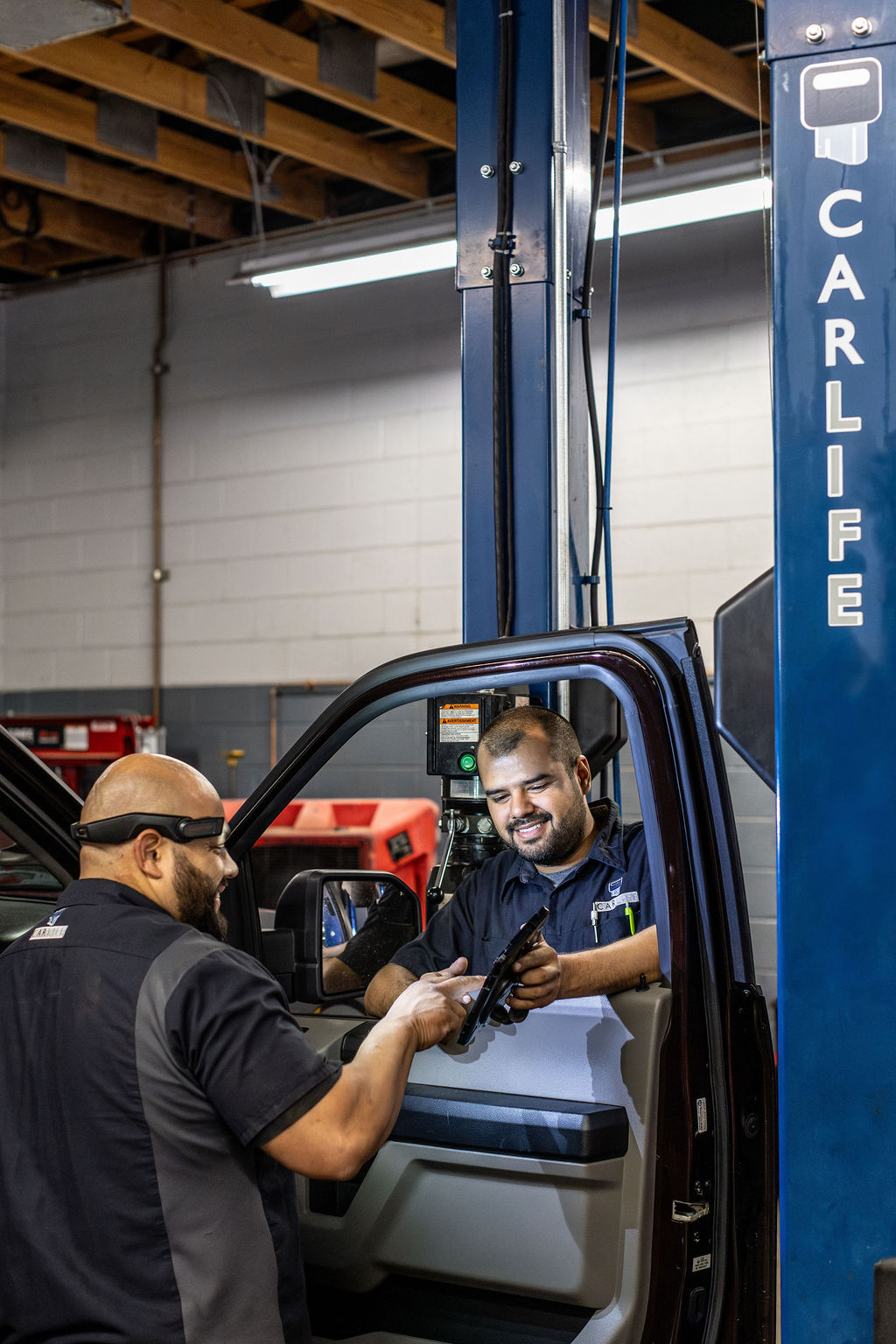 Two auto mechanics discussing a car part inside a vehicle at a workshop with a Carli Lift in the background.