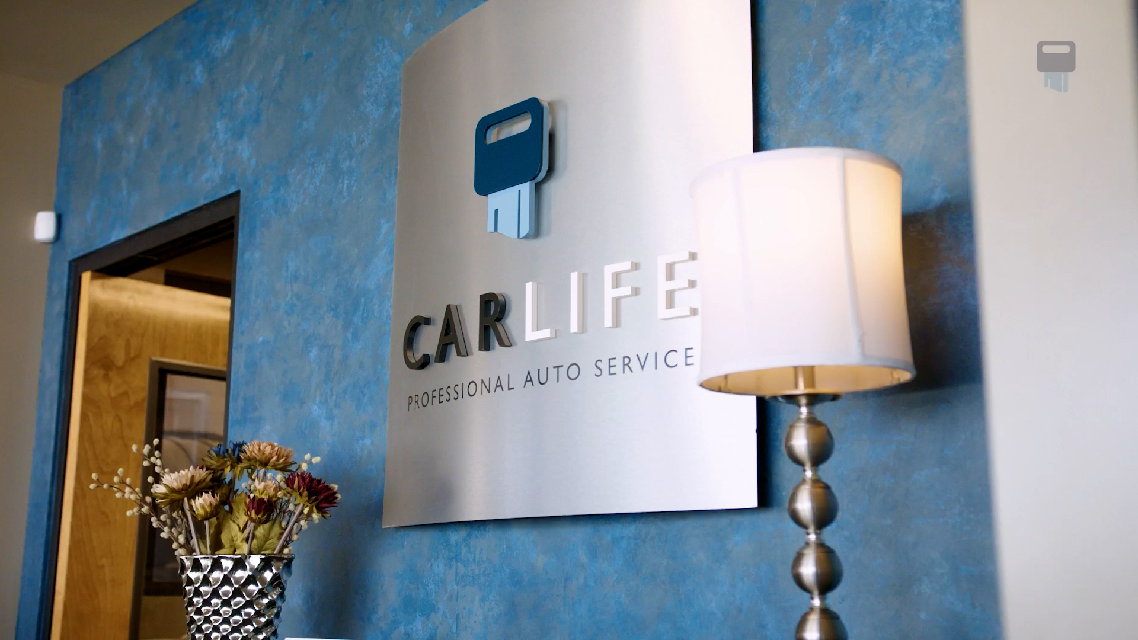 Wall sign displaying 'CARLIFE Professional Auto Service' with a key icon, next to a silver lamp and a vase with flowers.