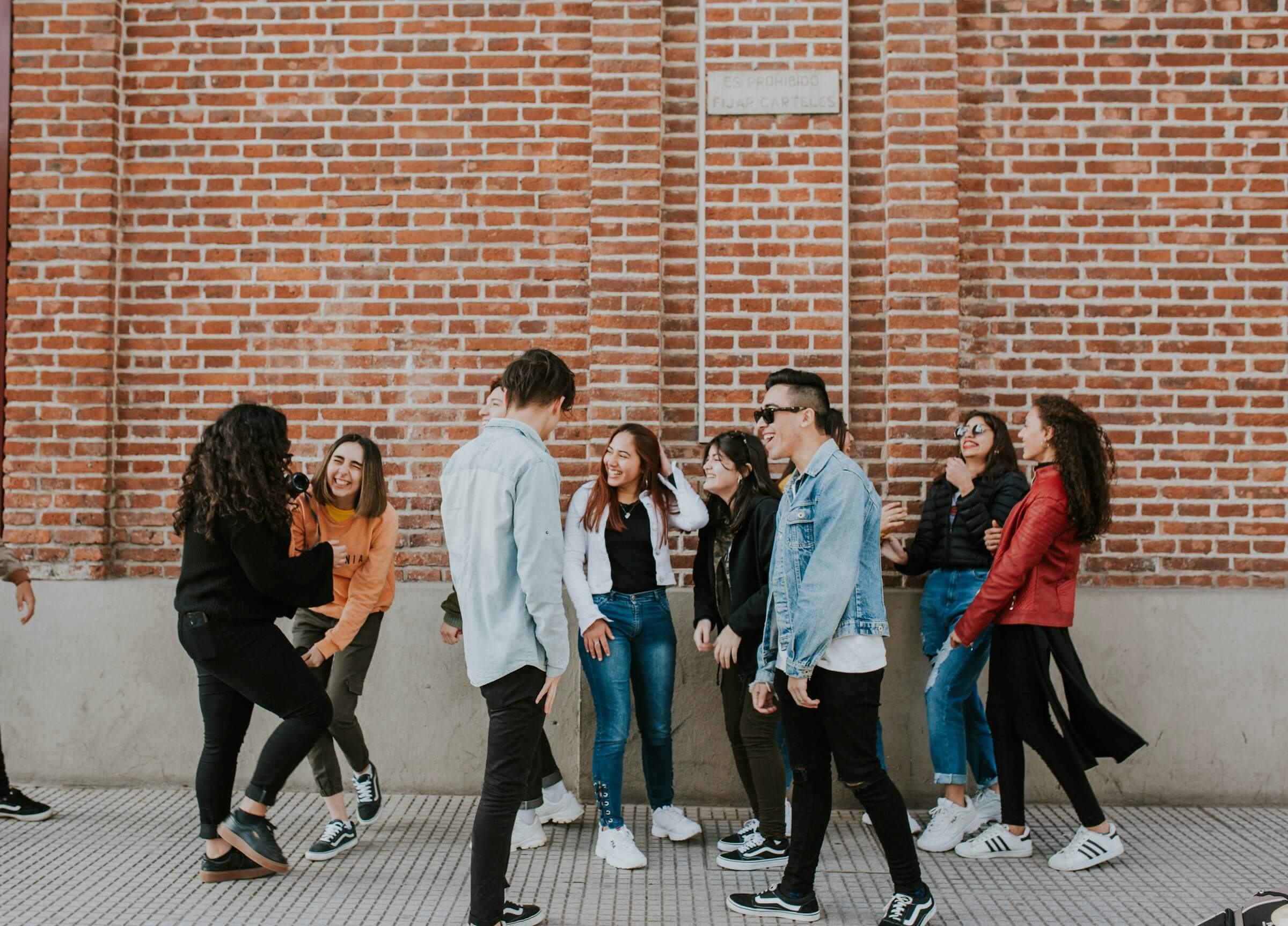 Group of young people laughing and chatting while standing on tiled pavement in front of a brick wall.
