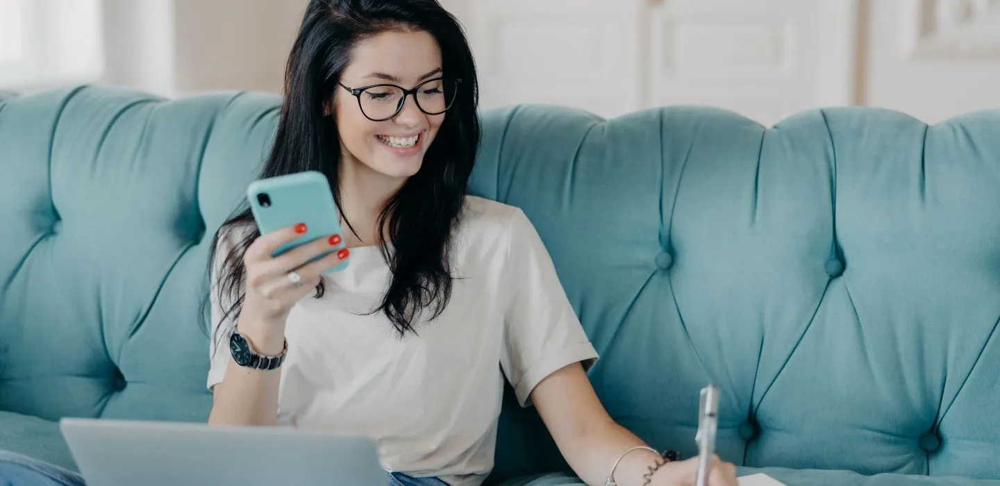 Junge Frau mit Brille sitzt lächelnd auf einem türkisfarbenen Sofa, hält ein Smartphone in der Hand und schreibt Notizen, vor ihr ein aufgeklappter Laptop.
