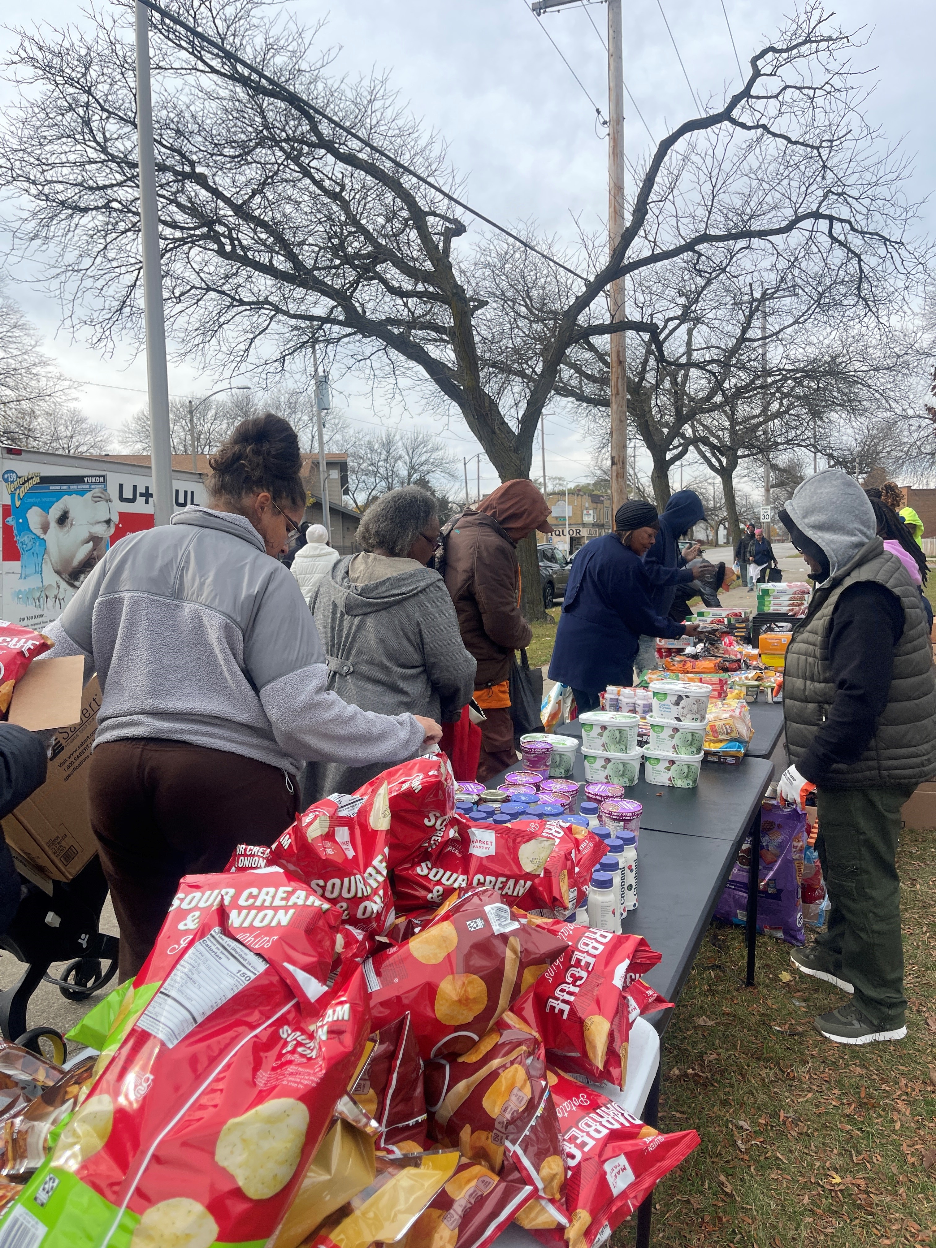 People lined up outdoors at a distribution table with snacks and groceries on a cloudy day.