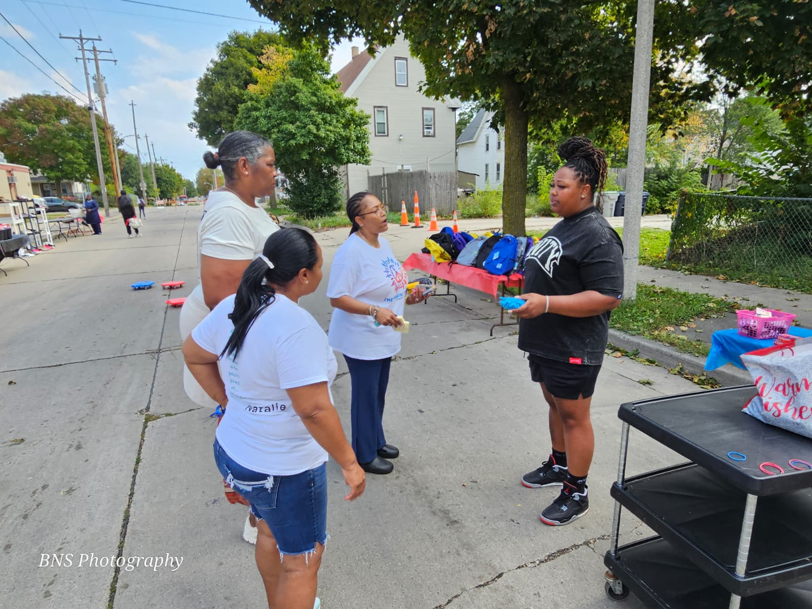 Four women standing on a neighborhood street engaged in conversation near tables with backpacks and supplies.
