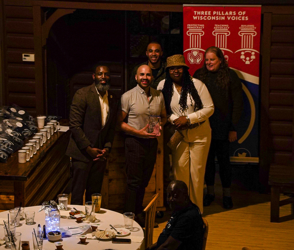 Group of five people posing indoors beside a table with drinks and food, in front of a red banner titled 'Three Pillars of Wisconsin Voices'.