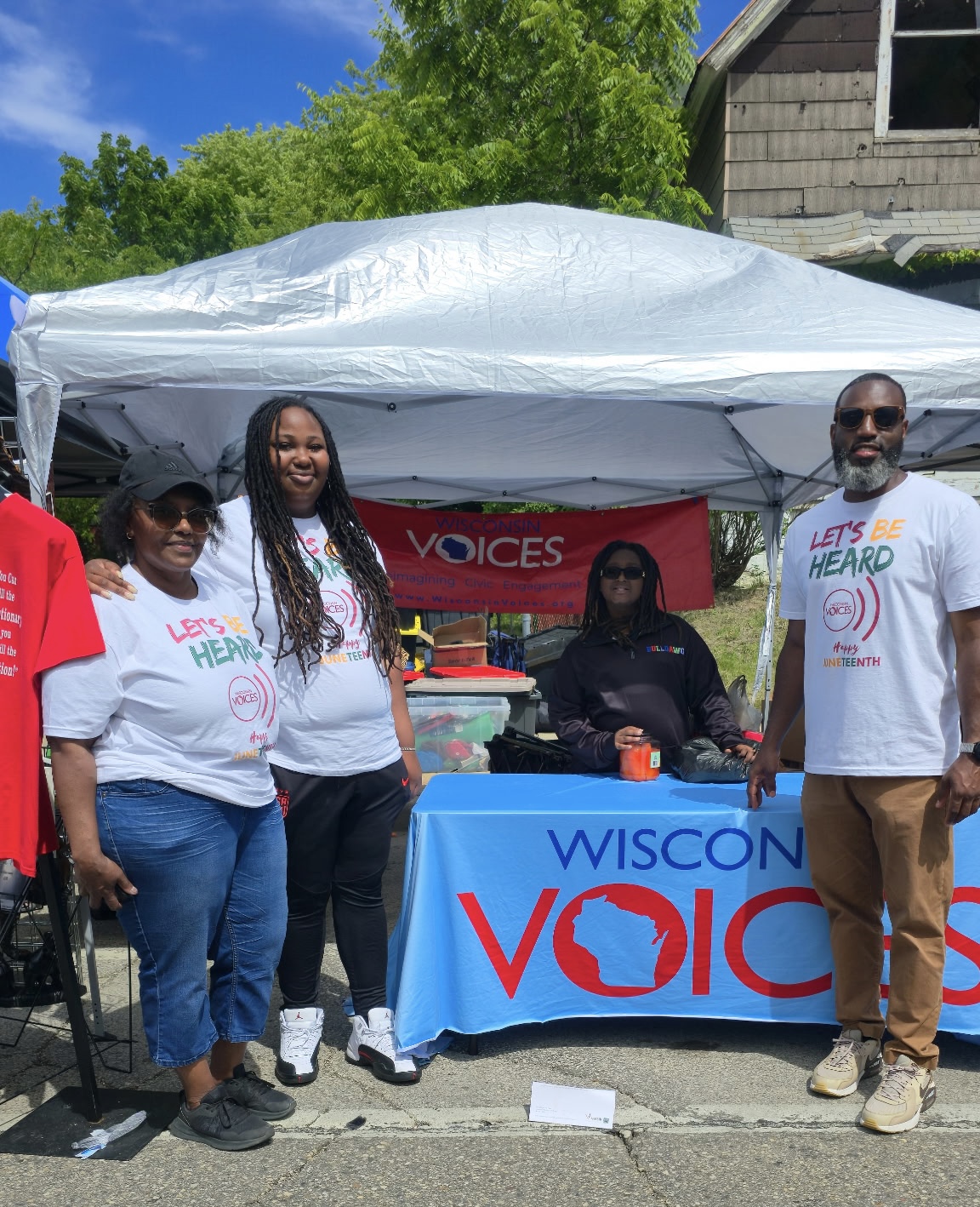 Group of five people posing indoors beside a table with drinks and food, in front of a red banner titled 'Three Pillars of Wisconsin Voices'.