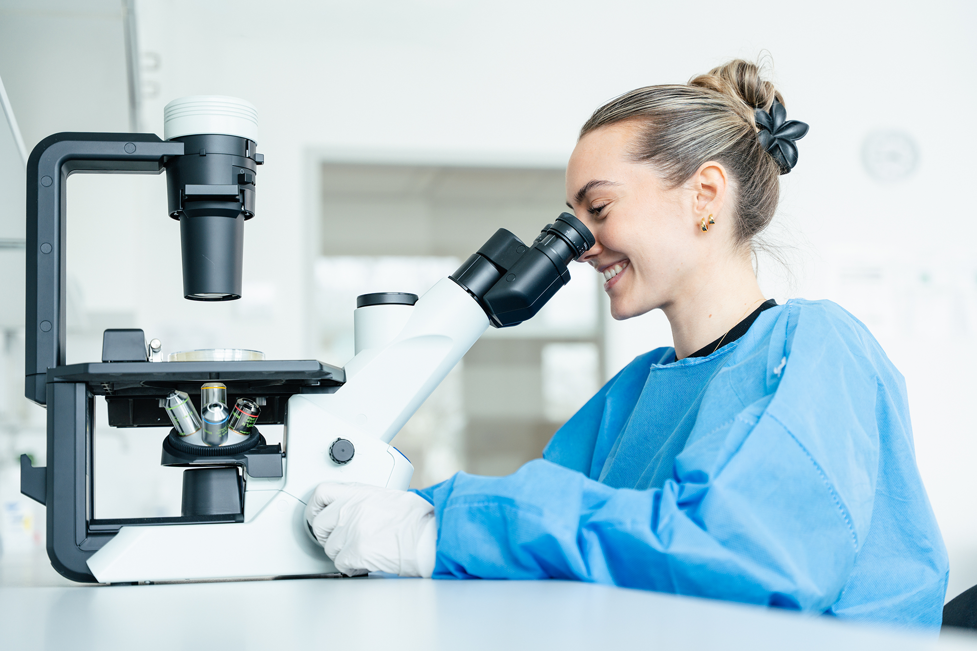 Smiling female scientist in blue lab coat looking through a white microscope in a bright lab.