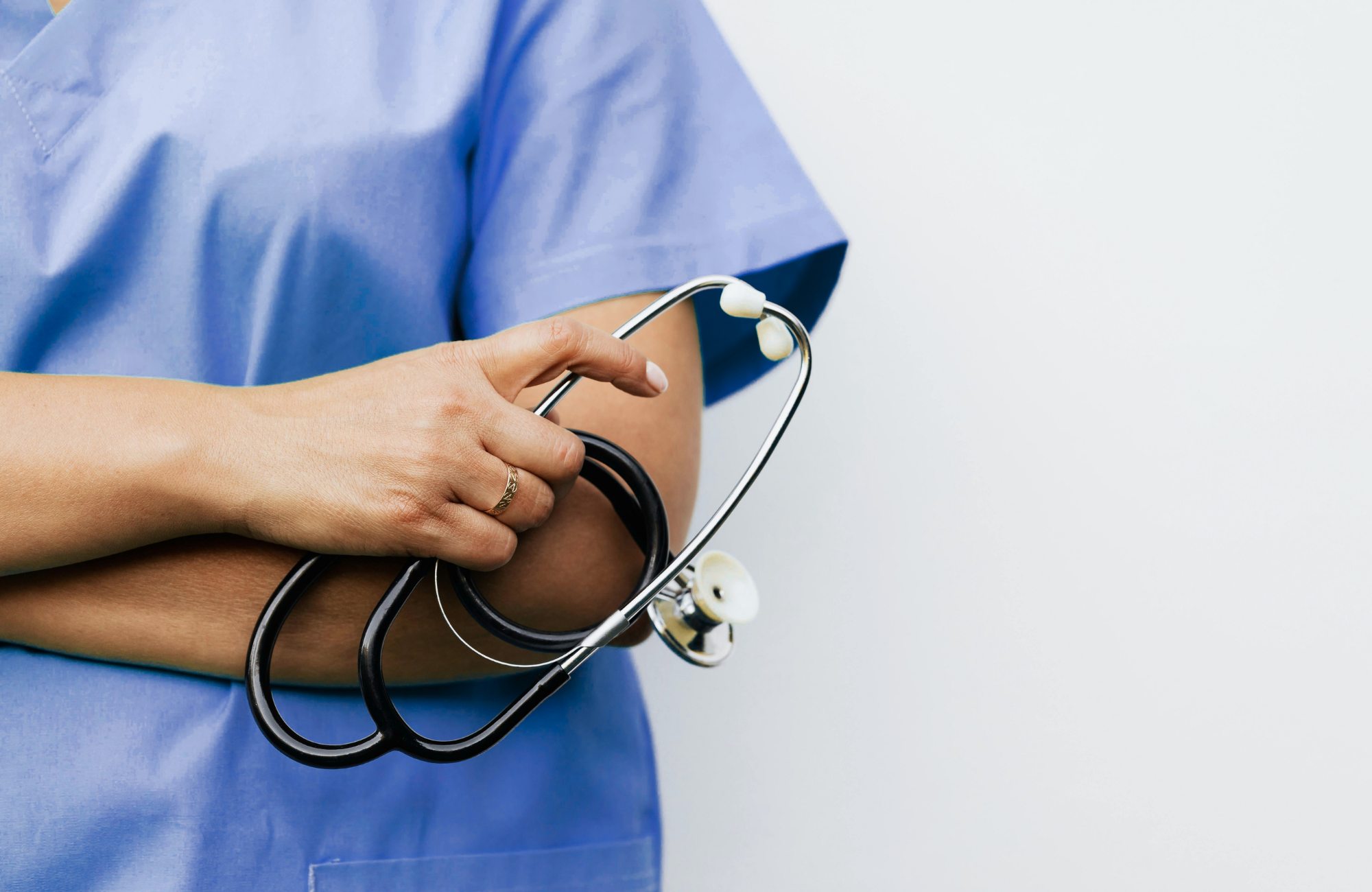 Medical professional in blue scrubs holding a stethoscope against a white background.