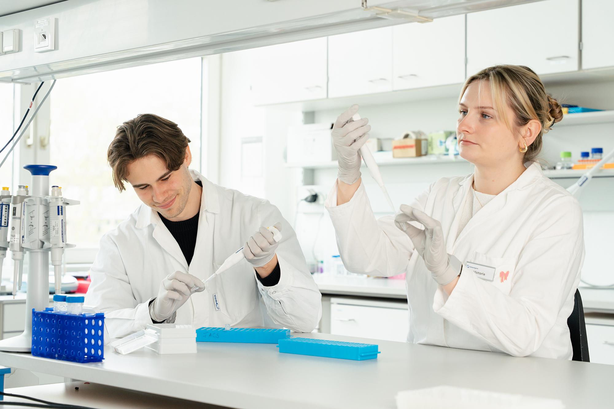 Two scientists in white coats working with pipettes and test tubes in a bright laboratory.