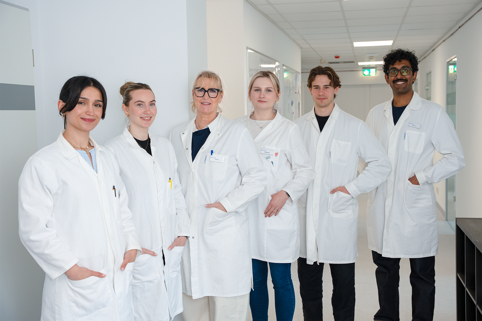 A team of six diverse healthcare lab scientists in white lab coats standing in a hospital hallway smiling at the camera.