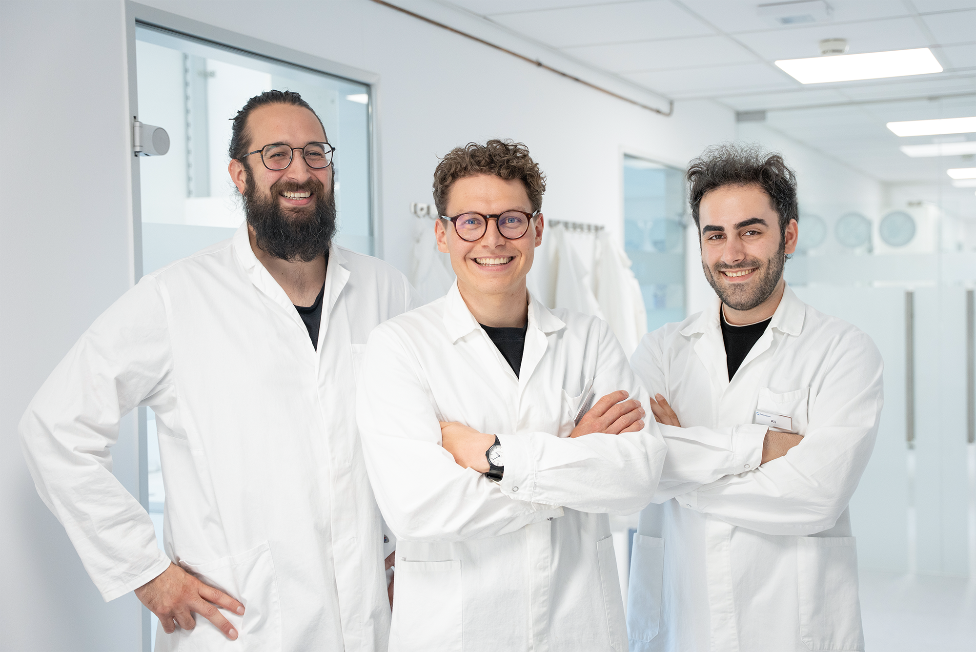 Three male tech scientists in white lab coats smiling inside a bright lab hallway.