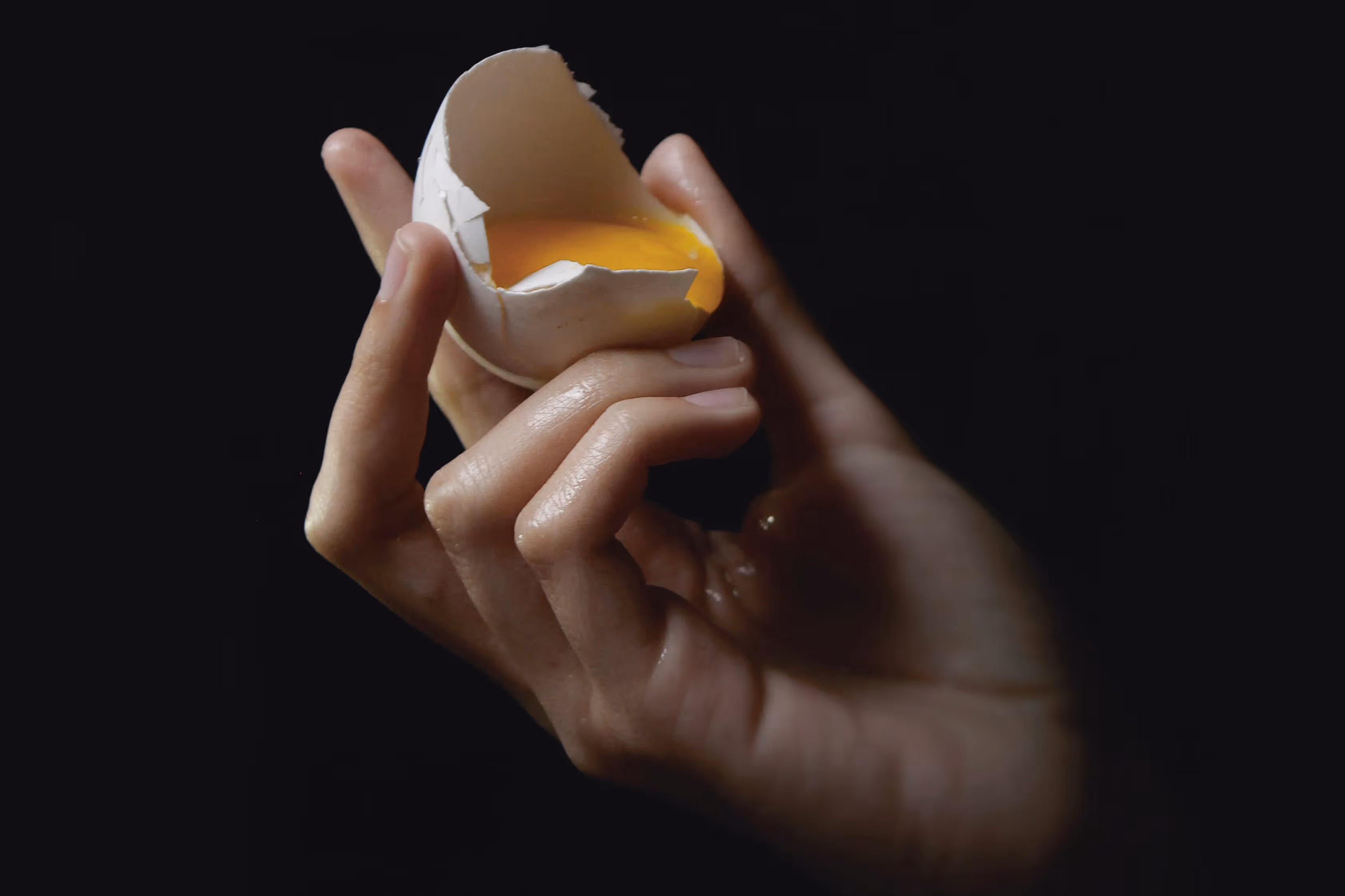 Hånd som holder en knekt eggeskall med eggeplommen synlig, mot mørk bakgrunn